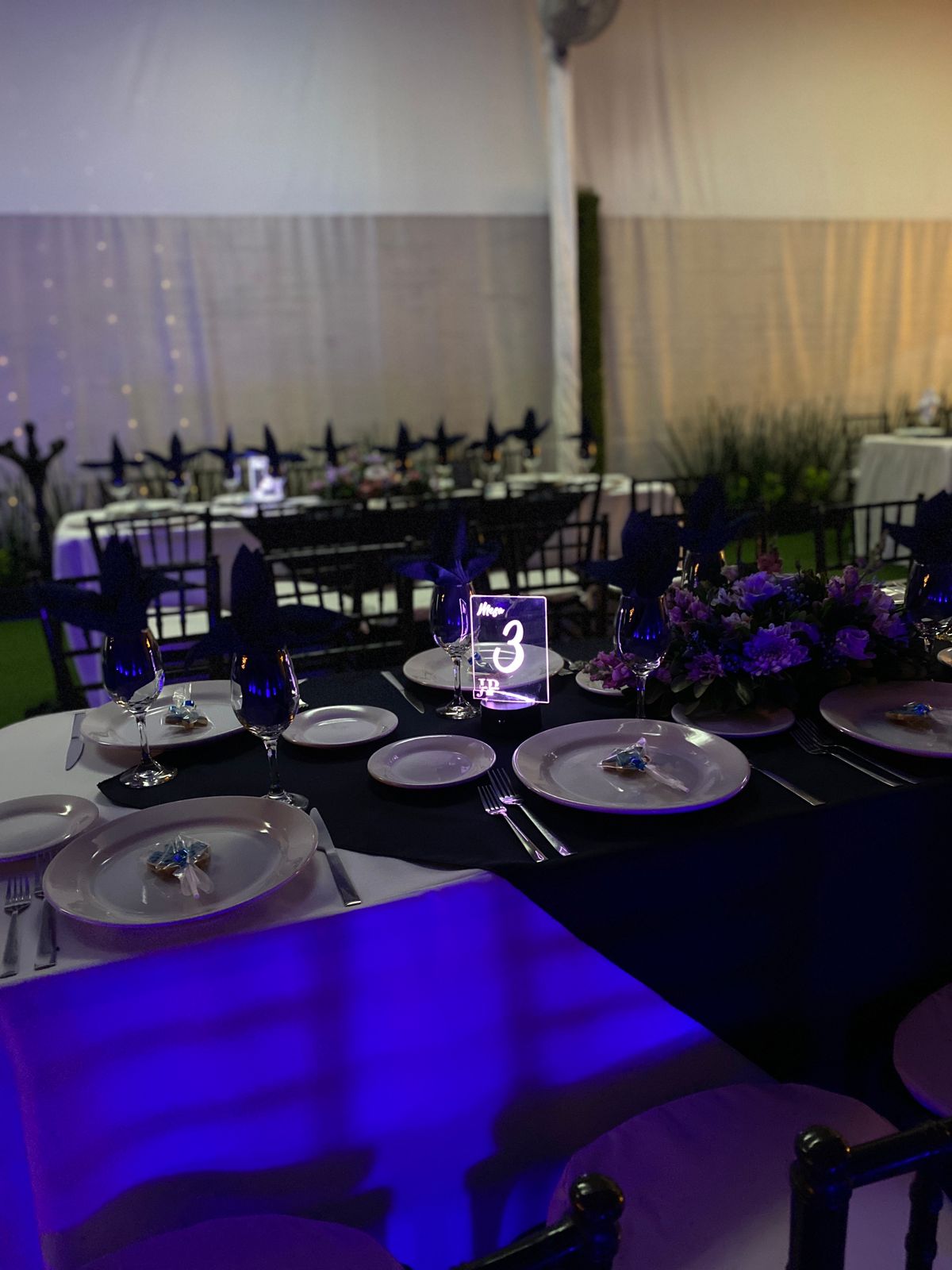 Mesa con mantel azul marino, número de mesa luminoso y centro floral morado bajo la carpa