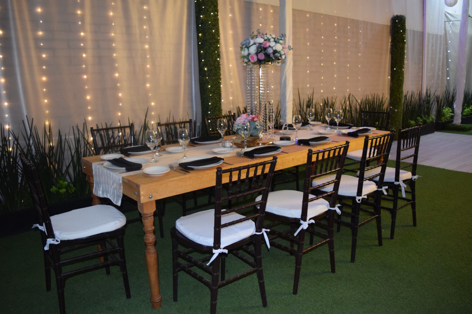 Mesa larga de madera con cortinas iluminadas, sillas negras y cojines blancos
