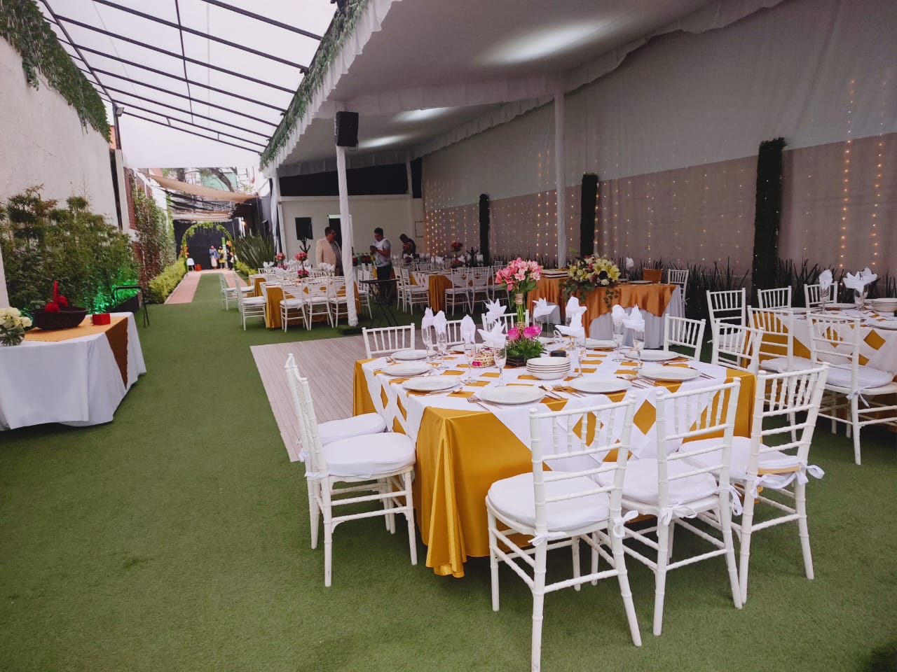 Terraza techada del jardín con mesas vestidas en blanco y dorado y corredor verde