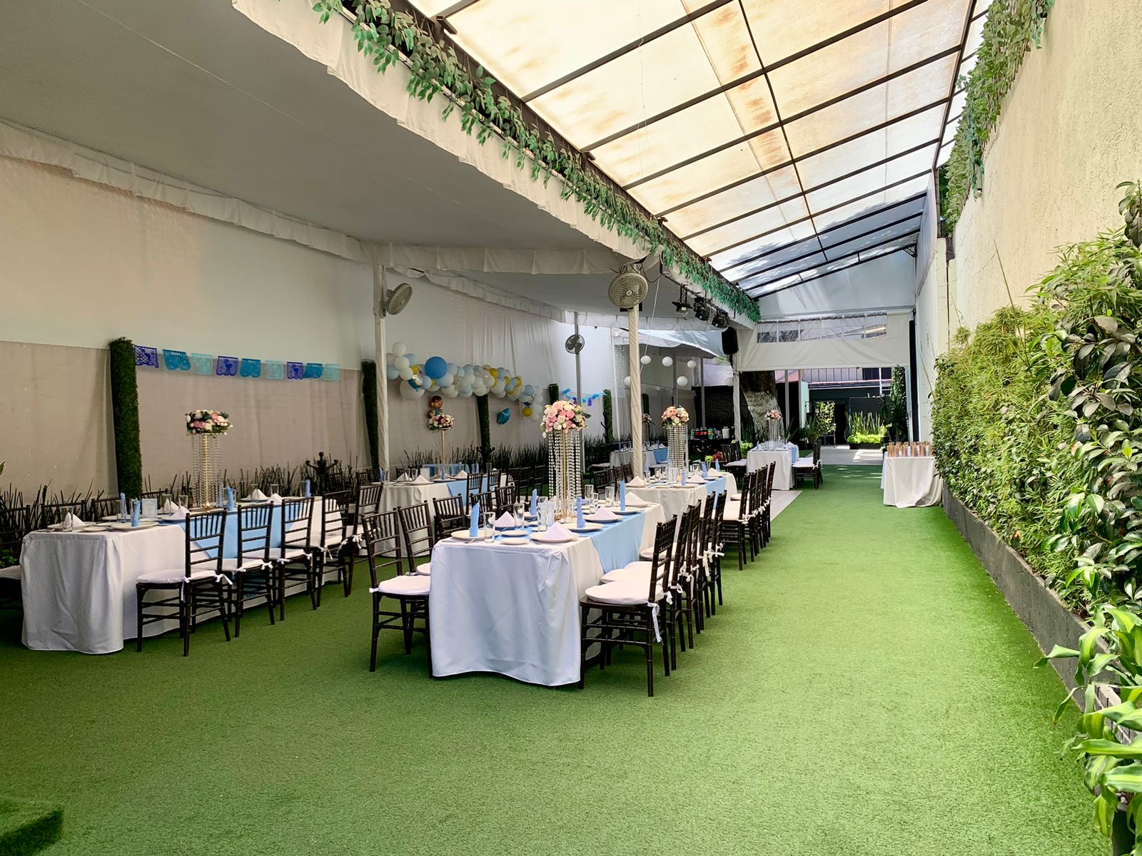 Cobertizo con corredor verde y mesas largas vestidas de blanco con servilletas azules