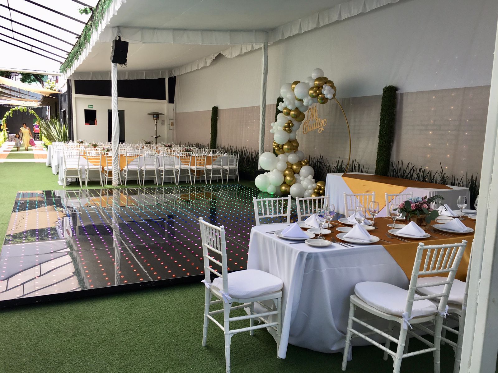 Pista de baile de LED iluminada junto a mesas con camino dorado y arco de globos blanco y dorado