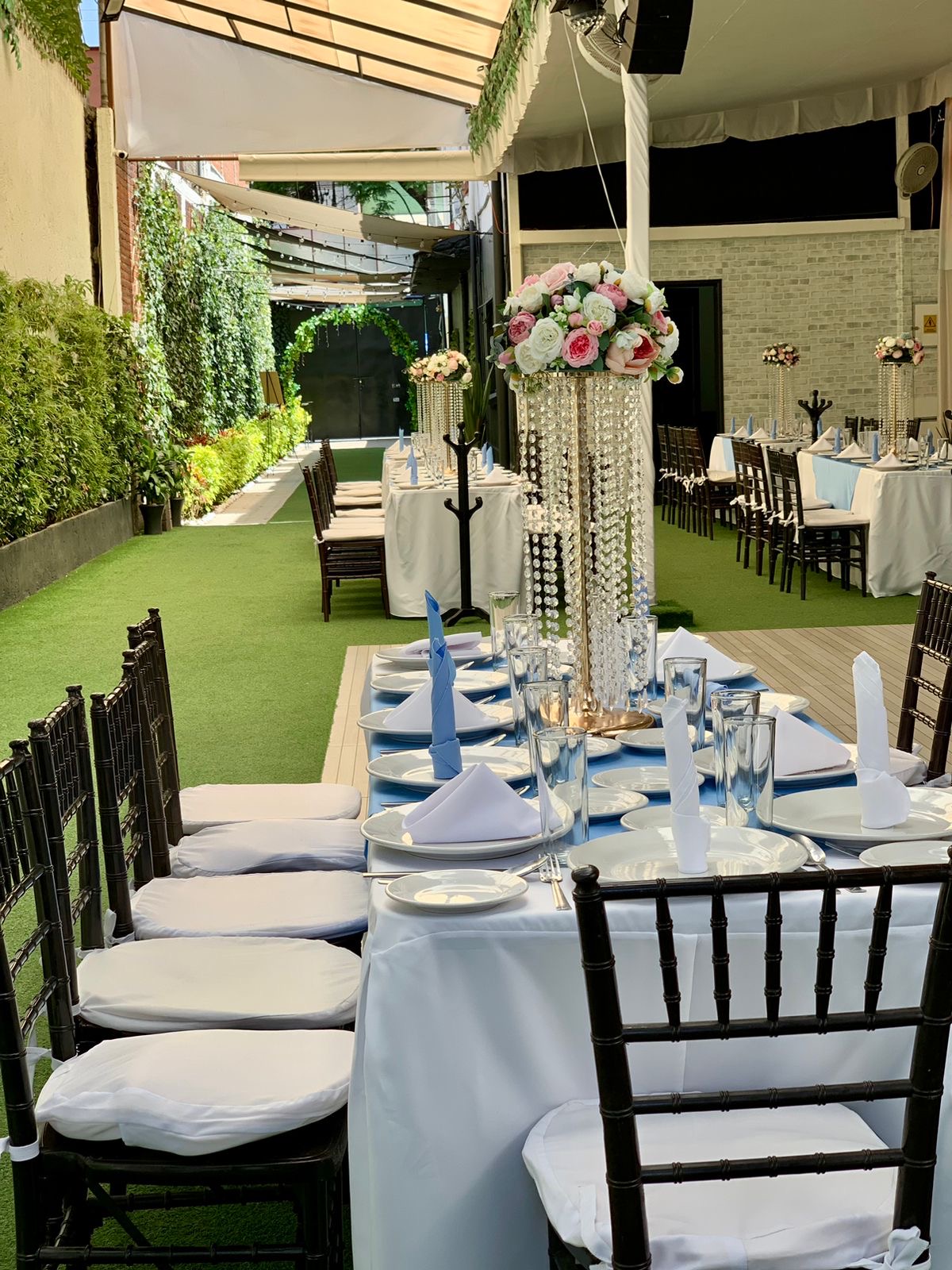 Mesa larga formal con sillas negras, mantel blanco, servilletas azules y centro de cristal con flores rosas