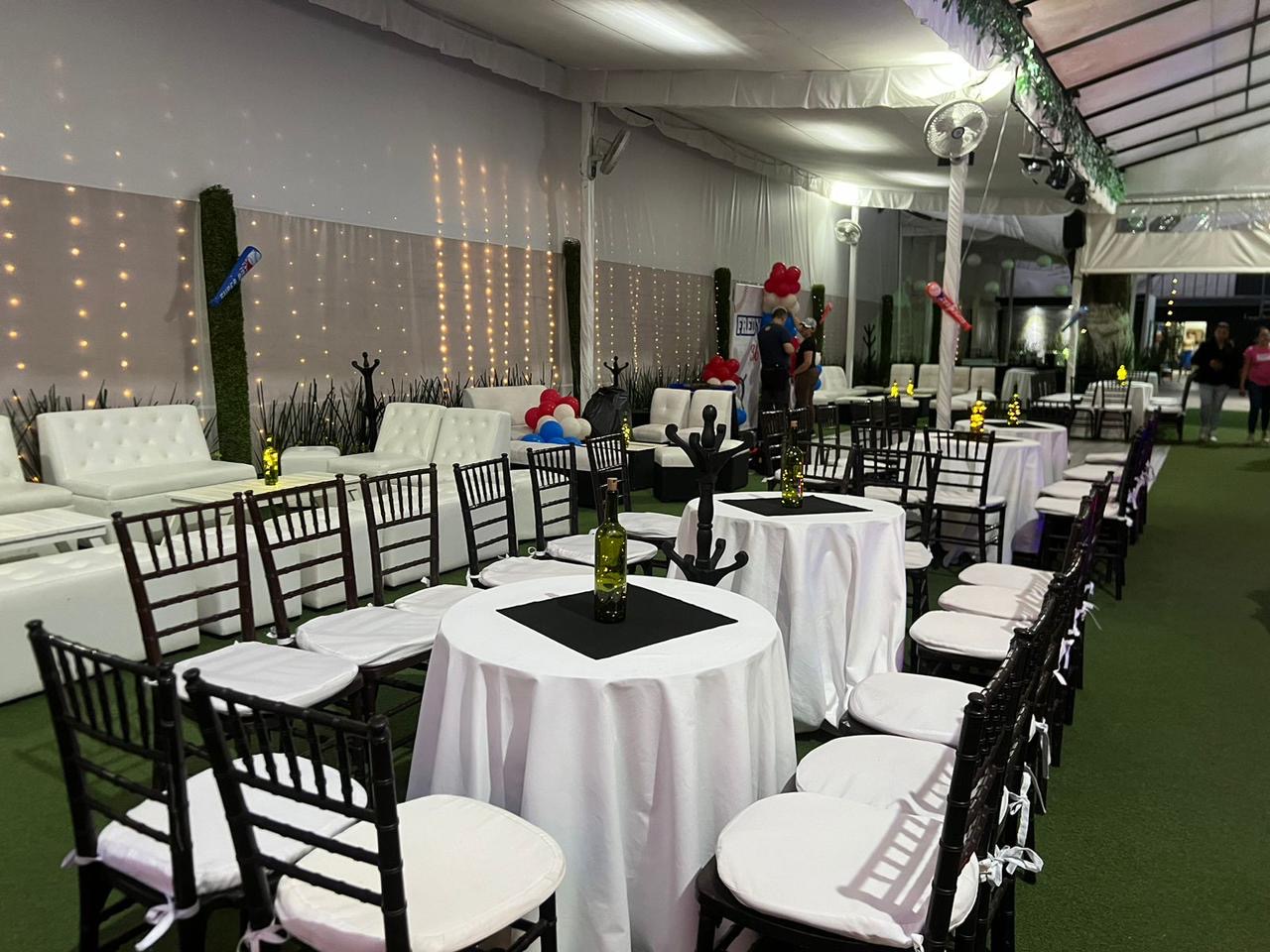 Zona lounge con salas blancas, mesas redondas de mantel blanco y sillas negras bajo la carpa