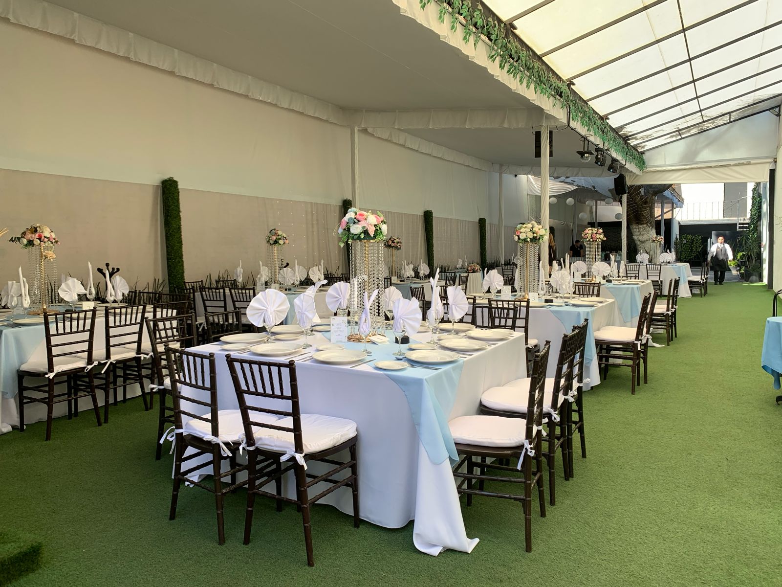 Carpa con mesas de mantel blanco, servilletas azules y centros de cristal con flores