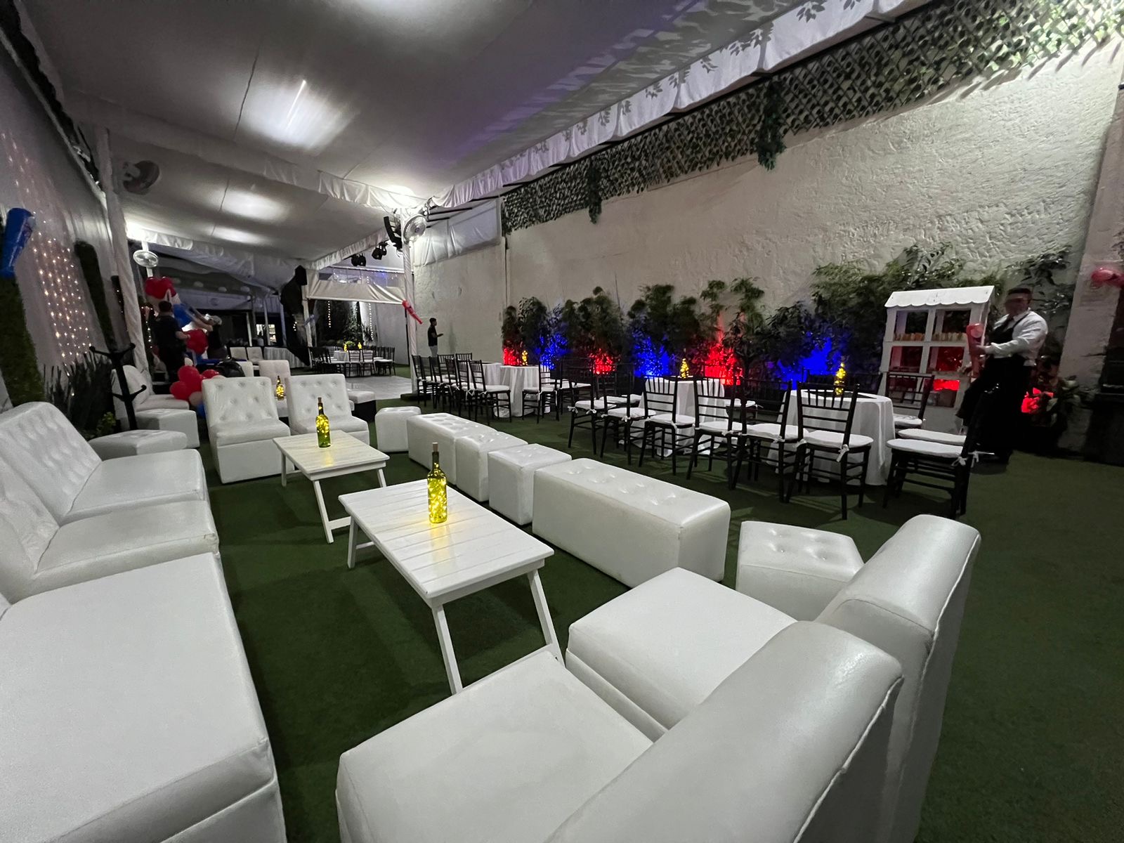 Salón lounge con salas blancas, cojines y vegetación con luces de colores