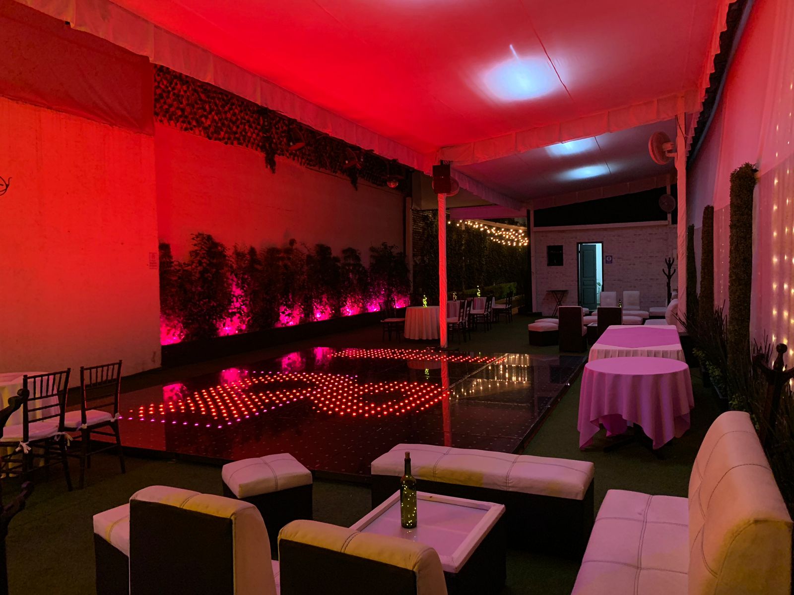 Pista de baile iluminada en rojo con zona lounge blanca y mesas de mantel rosa
