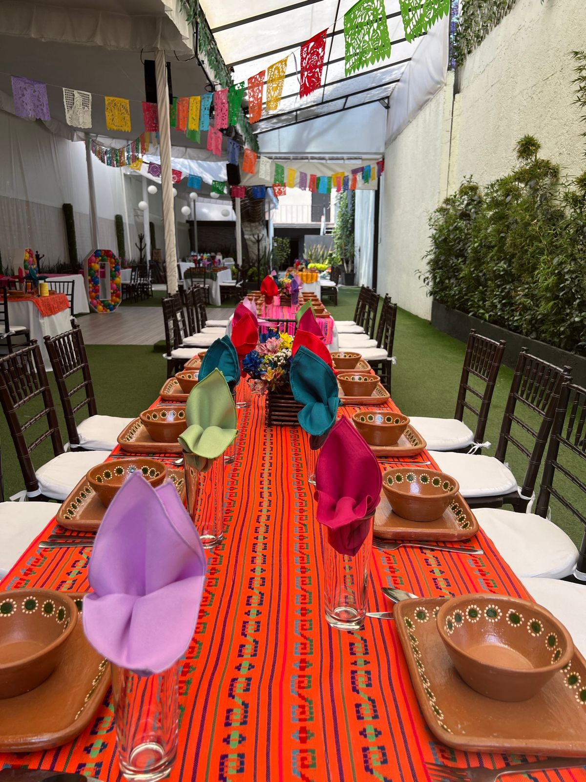 Corredor verde con mesa larga de mantel naranja mexicano, barro y papel picado