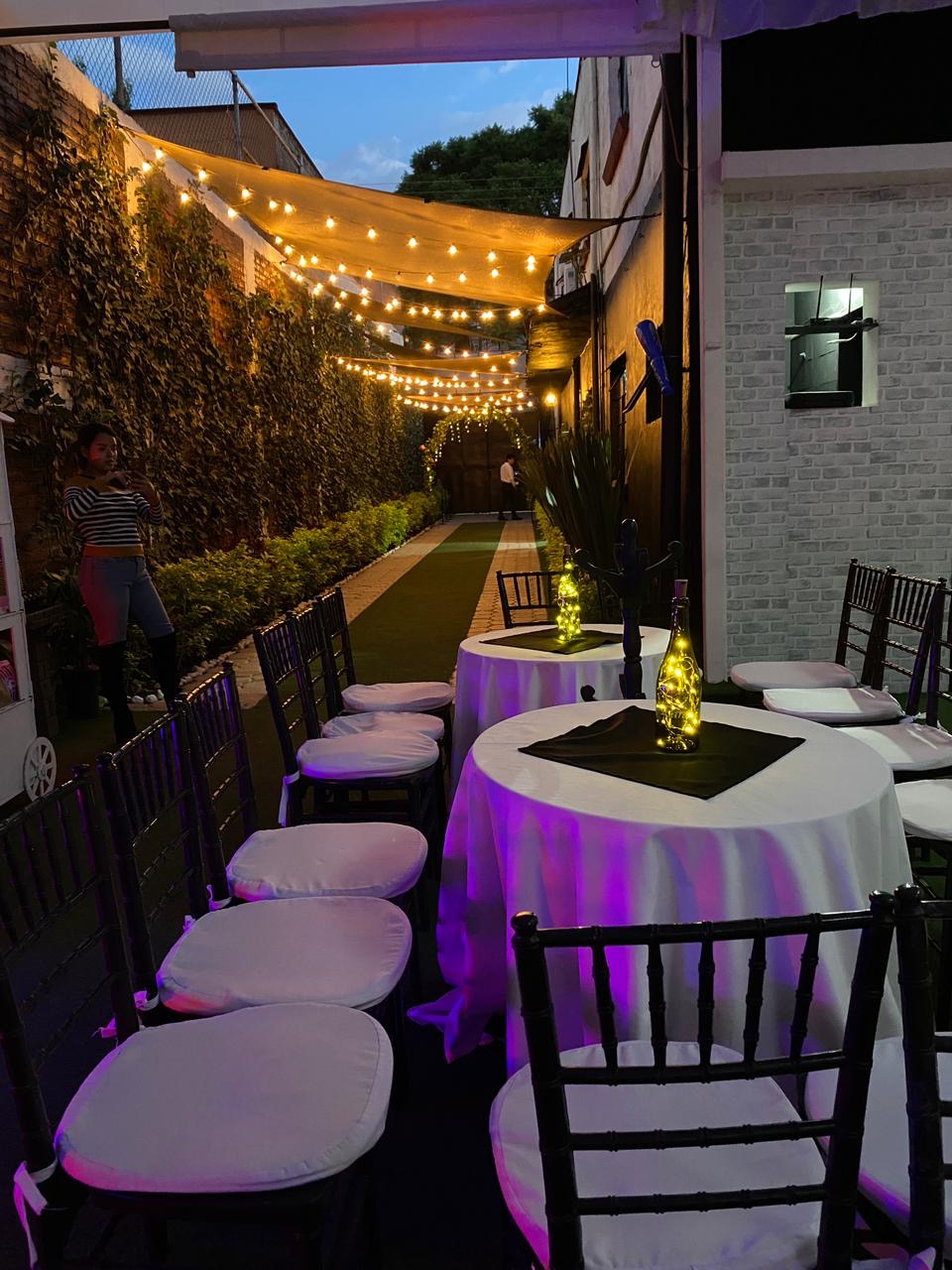Mesas redondas de lounge con mantel blanco, sillas negras y botellas luminosas bajo el túnel de luces
