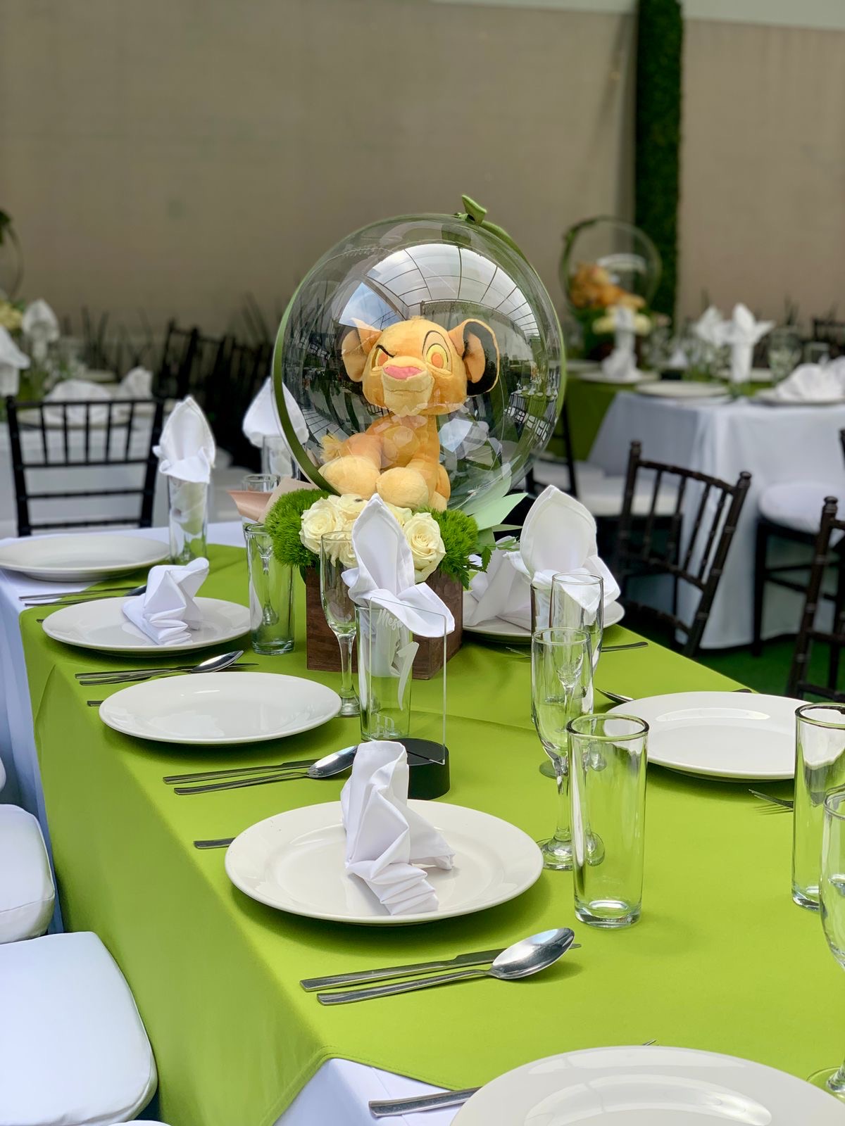 Mesa con mantel verde, centro de peluche en burbuja transparente y rosas blancas