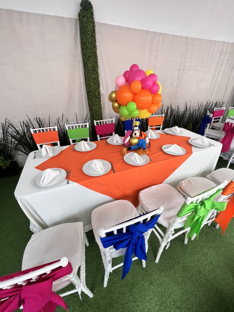 Mesa infantil larga con mantel naranja, sillas de colores y centro de globos multicolor